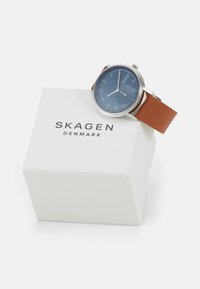Skagen-klocka med blå urtavla, hölje i silverton och brun läderrem, visad ovanpå en vit låda med "SKAGEN DENMARK" tryckt på.