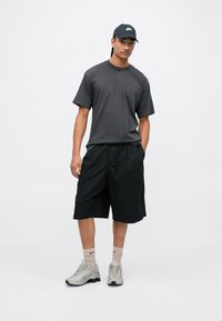 Camiseta gris de manga corta con logo en relieve, pantalones cortos negros hasta la rodilla, zapatillas deportivas grises y una gorra oscura. Conjunto de ropa deportiva informal.