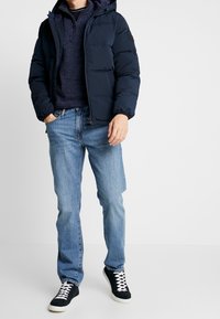 Marinblå puffjacka med huva, lättviktig, strukturerad tyg. I kombination med en mörk tröja, ljusblå jeans och svarta sneakers med vita snören.