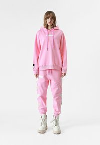 Sudadera de gran tamaño en color rosa con cordones y un parche con logo, combinada con pantalones de chándal ajustados a juego que presentan bordados laterales y costuras en contraste.