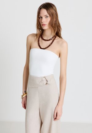Femme aux longs cheveux bruns portant un haut blanc sans bretelles, un pantalon beige taille haute avec ceinture, un collier de perles marron et des bracelets en or.