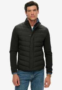 Doudoune noire avec un design léger, dotée d'un col haut, de boutons-pression et de panneaux texturés. Portée par-dessus une chemise en denim bleue.