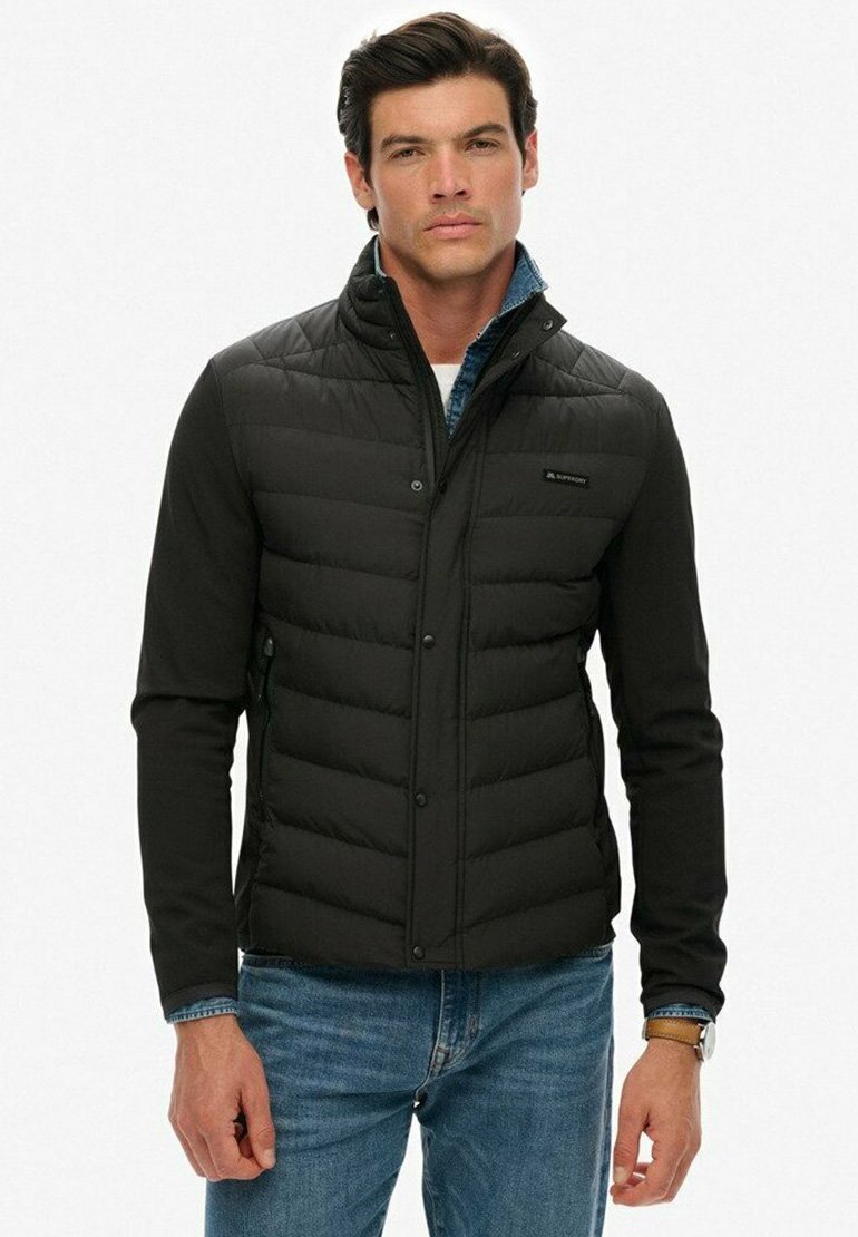Doudoune noire avec un design léger, dotée d'un col haut, de boutons-pression et de panneaux texturés. Portée par-dessus une chemise en denim bleue.