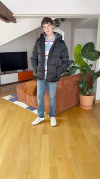 Μαύρο φουσκωτό μπουφάν με κουκούλα, φορεμένο πάνω από ένα ανοιχτό μπλε ριγέ πουκάμισο. Ανοιχτό μπλε τζιν και λευκά αθλητικά παπούτσια. Εσωτερικός χώρος με φυτά.