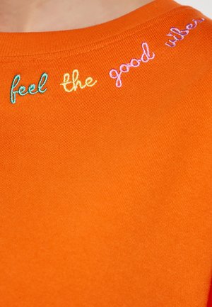 Oranje katoenen t-shirt met korte mouwen, voorzien van multikleurige borduursels die "voel de goede vibes" langs de halslijn weergeven.