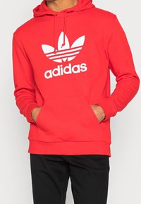Röd Adidas hoodie med en stor vit trefoil-logotyp. Har en framficka och snörning i huvan. Tillverkad av ett slätt material.