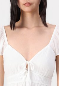 Vit klänning med korta puffärmar, en djup V-ringning och en knytfram. Tyget är lätt och har en subtil textur.