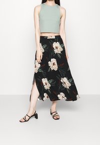 Falda maxi negra con patrones de peonías en beige y verde, combinada con un top corto ajustado de color verde claro. Presenta una abertura lateral y una tela suave.