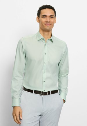 Mann steht, trägt ein hellgrünes Hemd mit Knöpfen, hellgraue Hose und einen dunklen Gürtel, mit einer Hand in der Tasche.