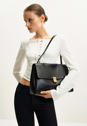 Femme aux cheveux tirés en arrière porte un haut blanc boutonné et un pantalon noir, tenant un sac à main en cuir noir avec une fermeture dorée.