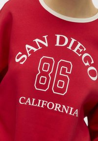 Sweatshirt rouge avec un col rond blanc. Présente un texte imprimé : "SAN DIEGO", "86", et "CALIFORNIE" en lettres grasses et contrastées.