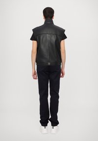 Μαύρο δερμάτινο γιλέκο χωρίς μανίκια με ψηλό γιακά, που διαθέτει κούμπωμα στη μέση, σε συνδυασμό με σκούρα παντελόνια και λευκά αθλητικά παπούτσια.