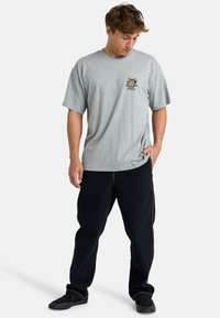 Gray cotton t-shirt with short sleeves and a crew neck. Features a colorful graphic on the left chest. Paired with black, loose-fitting pants.