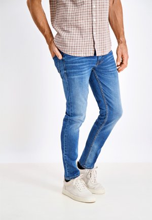Jeans in denim blu slim-fit con leggeri sbiadimenti, abbinati a sneakers beige chiaro. La camicia presenta un motivo a quadri marrone e bianco.