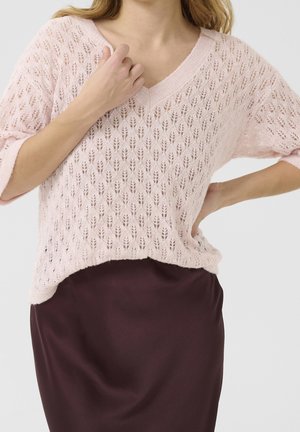 Kvinde iført en lyserød blondestrikket V-hals sweater og en mørkelilla nederdel, med den ene hånd, der rører ved halsudskæringen og den anden på hoften.