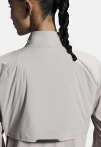 Chaqueta deportiva de color gris claro con un cuello alto, textura suave y un diseño trasero en capas. Presenta costuras cosidas y un aspecto minimalista.