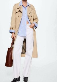 Beige trenchcoat over en blåstribet skjorte, hvide vide bukser og sorte ankelstøvler. Brun tote taske med en glat overflade.