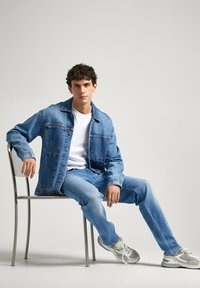 Chaqueta de denim y jeans en azul claro, camiseta blanca debajo, sentado en una silla de metal, con zapatillas grises y un fondo neutro.