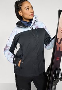 Melna un gaiši zila raiba slēpošanas jaka ar rāvējslēdzēju priekšpusē, regulējamu kapuci un vairākām kabatām; valkā persona, kas tur slēpes.