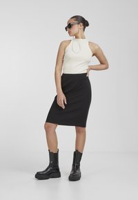 Falda negra acanalada hasta la rodilla, top sin mangas crema y botines negros gruesos hasta el tobillo. El conjunto se complementa con unas gafas de sol negras de gran tamaño.