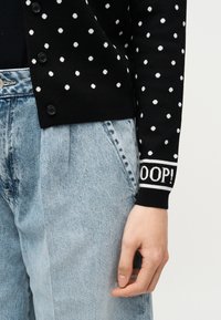 Μαύρο ζακέτα με λευκές πουά, κείμενο "OOOP!" στη μανσέτα, συνδυασμένο με ανοιχτό μπλε τζιν ψηλής μέσης, δεξί χέρι χαλαρό δίπλα στο σώμα.