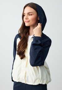 Lichte jas, gekleurd in navy en crème, met capuchon, elastische manchetten en een ritssluiting. Glad oppervlakte, casual ontwerp.