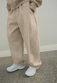 Béžové tepláky vyrobené z mäkkého materiálu, s voľným strihom a bočnými vreckami, skombinované s bielymi teniskami na textúrovanom povrchu.