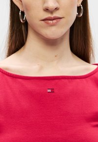 Rdeča bombažna majica z širokim izrezom. Ima majhen, vzorčen okras v rdeči, beli in modri barvi blizu ovratnika. Gladka tekstura.