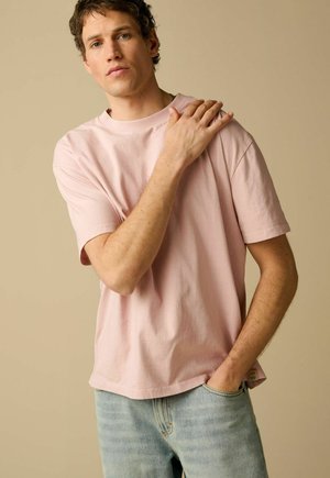 Junger Mann in hellrosa T-Shirt und hellblauen Jeans, Hand auf der Schulter, neutraler Hintergrund.