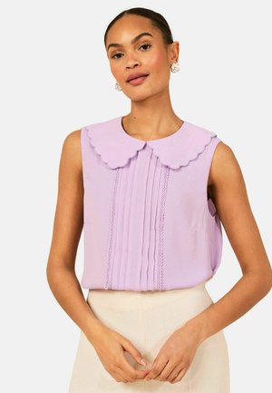 Žena v lila blůze bez rukávů s vlnitým límečkem Peter Pan a vertikálními plisovanými prvky, ve spojení s béžovými kalhotami s vysokým pasem.