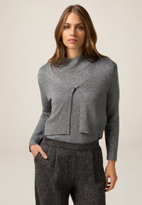 Maglione grigio corto con collo alto, lavorazione a maglia testurizzata e dettaglio in vita con nodo. Indossato con pantaloni grigio scuro a fantasia.