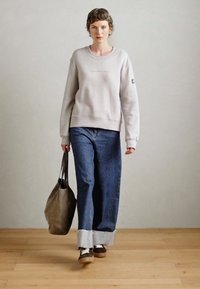 Lysegrå hettegenser med rund hals og broderidetaljer, kombinert med vide mørke jeans med frynset kant og brune sneakers.