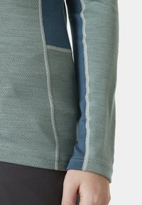 Długi rękaw top w zielono-niebieskim kolorze z teksturowanej tkaniny, kontrastującymi szwami po bokach oraz kieszenią na piersi.