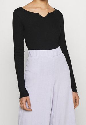 Person iført en sort langærmet tætsiddende top med bølget halsudskæring og højttaljede, lyse lilla brede bukser.