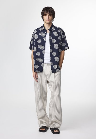 Camicia blu navy a maniche corte con stampe di conchiglie bianche, canotta bianca sotto, pantaloni a righe beige e sandali neri a ciabatta.