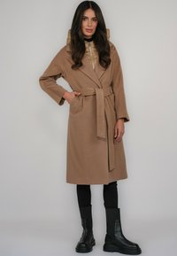 Płaszcz o kolorze wielbłądzim z paskiem do wiązania, szerokimi rękawami i naszywanymi kieszeniami, nałożony na pikowaną złotą kamizelkę i czarne legginsy. Grube czarne botki.