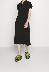 Black button-up dress with short sleeves, reaching mid-calf. Paired with green and black striped fuzzy slip-on shoes. Smooth texture.