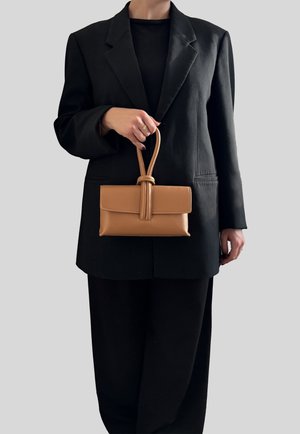Persona con blazer e pantaloni neri che tiene una piccola borsa in pelle color cuoio con manico annodato davanti a uno sfondo grigio uniforme.