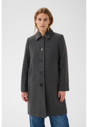 Femme portant un manteau boutonné gris foncé longueur genou avec deux poches latérales sur un jean bleu, sur fond uni.