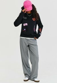 Čierna mikina s farebnou výšivkou, sivé nohavice s prúžkami a ružová čiapka. Biely športový obuv dodáva kontrast.