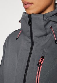 Gros plan d'une personne portant une veste grise à col haut avec des zips et des cordons roses, lèvres et menton visibles sur fond blanc.