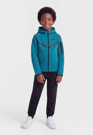 Smilende dreng iført teal Nike lynlås-hættetrøje, sorte bukser og hvide sneakers, der står mod en ensfarvet hvid baggrund.