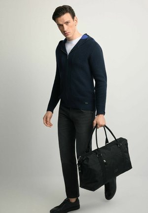 Granatowa bluza z zamkiem i teksturowanym wzorem, czarne dżinsy oraz czarna torba sportowa. Sneakersy mają gładkie wykończenie z minimalnymi zdobieniami.