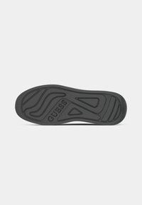 Semelle de chaussure en caoutchouc noire avec des motifs texturés ondulés et triangulaires, et le nom de la marque "GUESS" embossé au centre.