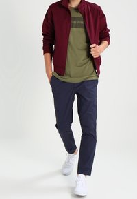 Chaqueta bomber burdeos sobre una camiseta verde oliva con un gráfico oscuro, combinada con pantalones azul marino y zapatillas blancas. Texturas de tejido suaves.