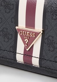 Borsa nera in tessuto con una texture, dotata di un logo triangolare dorato e strisce bordeaux e bianche. Il materiale presenta un motivo sottile e dettagli cuciti.