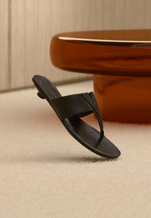 Sort læder tåsandal med lav hæl, svævende over beige tæppe nær et rundt brunt bord i en minimalistisk indendørs indstilling.