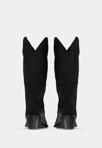 Ζεύγος μαύρων καουμπόικων μποτών μέσου μήκους με συσσωρευμένα τακούνια και διακοσμητική ραφή, εμφανιζόμενες από πίσω σε απλό φωτεινό φόντο.