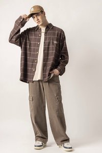 Giovane uomo che indossa una camicia button-up a quadri marrone, pantaloni cargo beige, un berretto beige e sneakers, che aggiusta il berretto con una mano in un ambiente neutro.