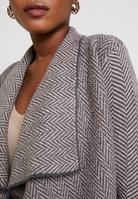 Šedý šál s vzorem rybí kosti a šálovým límcem. Měkká textura, otevřený přední díl a kontrastní švy podél okrajů.